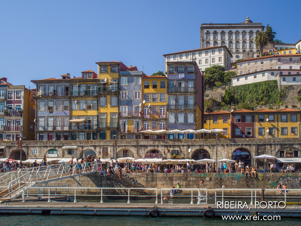 In this photograph we can see the historic riverside promenade of the Ribeira do Porto, with the wall, the restored houses on the hillside and the Paço Episcopal do Porto at the top of the hill